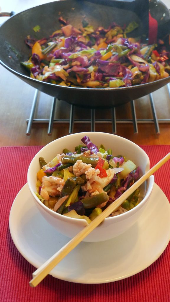 Wok mit Gemüse in Sauce im Hintergrund; im Vordergrund eine Schale mit Stäbchen, Reis und Wokgemüse