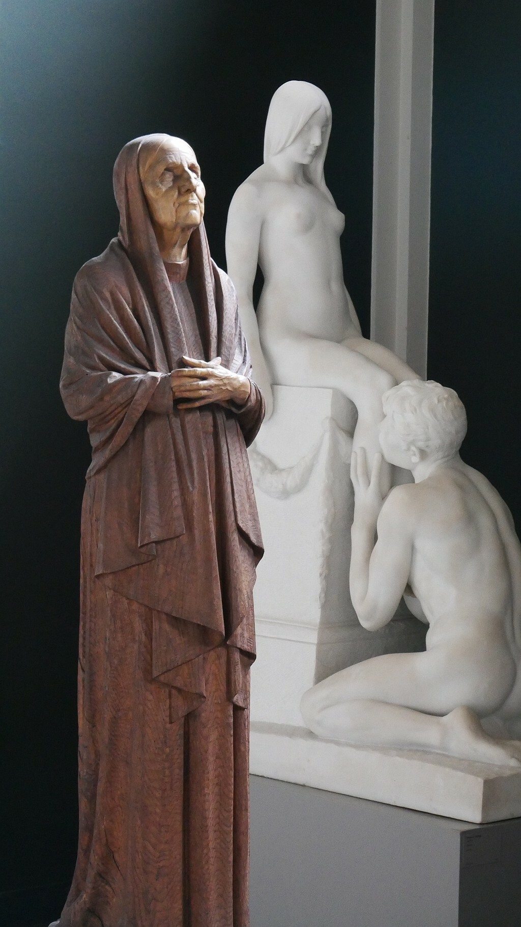 sculpture of old woman with folded hands in the foreground. sculpture of nude couple in the background.